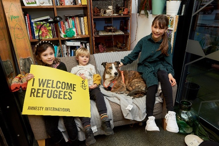 Three children sitting on a couch with a dog holding a sign which reads I welcome refugees